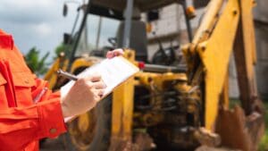 A mechanical engineer is using checklist form to verify the excavator or earthmover machine (as background) condition before operating at construction site. Industrial working scene.